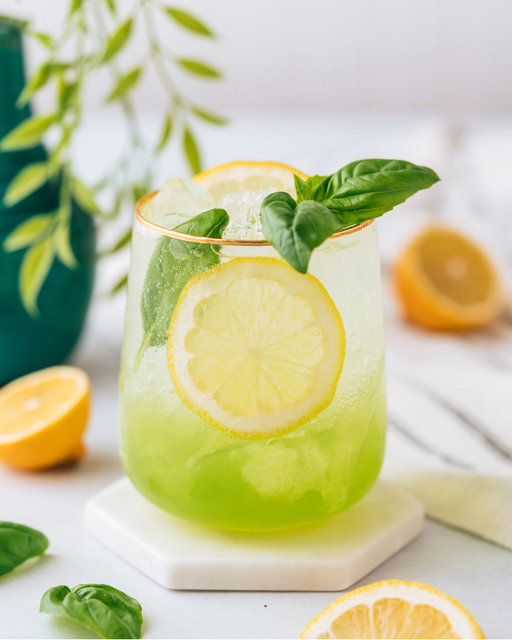 Basil Limoncello Cocktail in glass with ice, lemon wheel, and basil sprig with cut lemons and basil in background