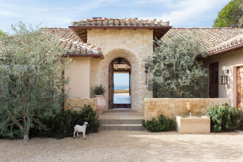 Mediterranean-style beach home exterior with pristine curb appeal featuring a fountain, tidy landscaping, a sheep figure, and slice of an ocean view through door