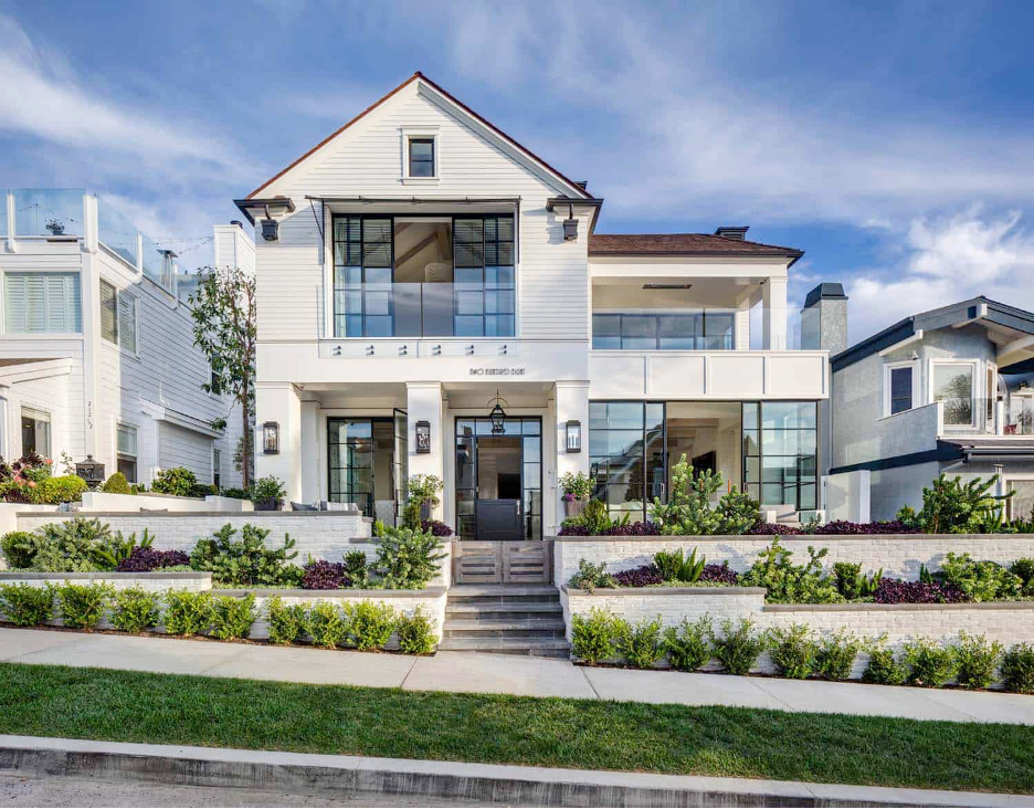 Curb appeal of Corona Del Mar, California beach home with steps up past beautiful tiered white-brick gardn wall, green grass, expansive glass windows, exterior lighting, and large Dutch door