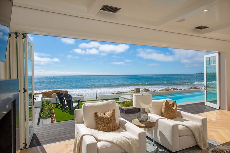 gorgeous wall of folding windowed doors opening onto views of the Pacific Ocean, an outdoor pool area, green lawn seating area of beach home 