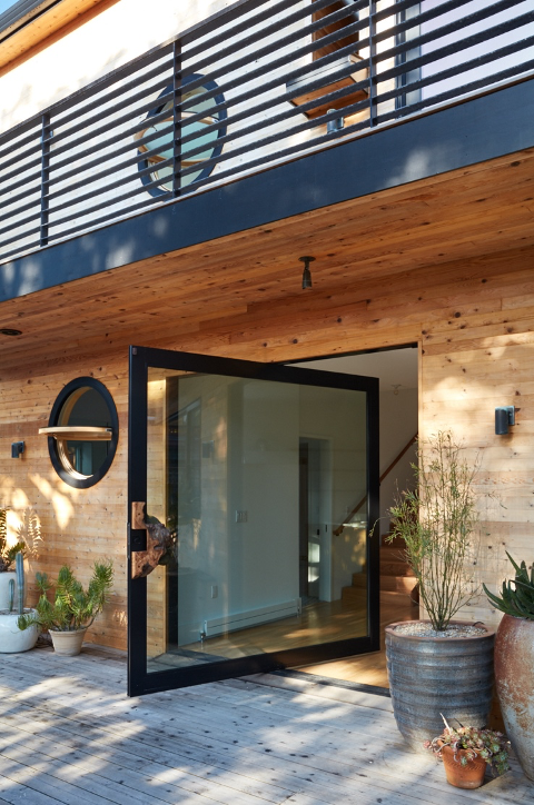 Pivot door with live-edge walnut handle and porthole-style window in backyard entrance to San Francisco surf pad