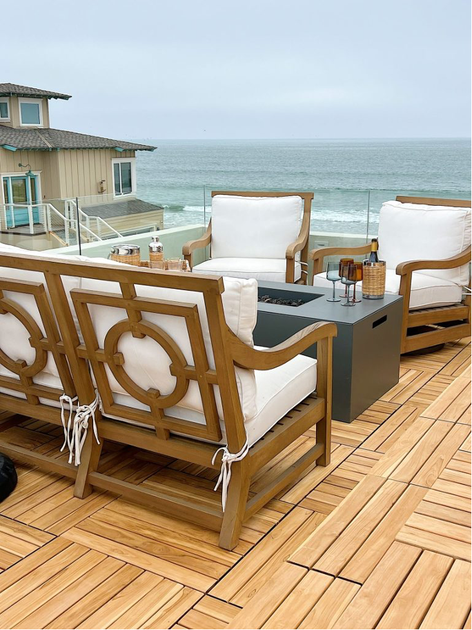 Seaside balcony with teak outdoor furniture, white cushions, and natural gas fire pit.