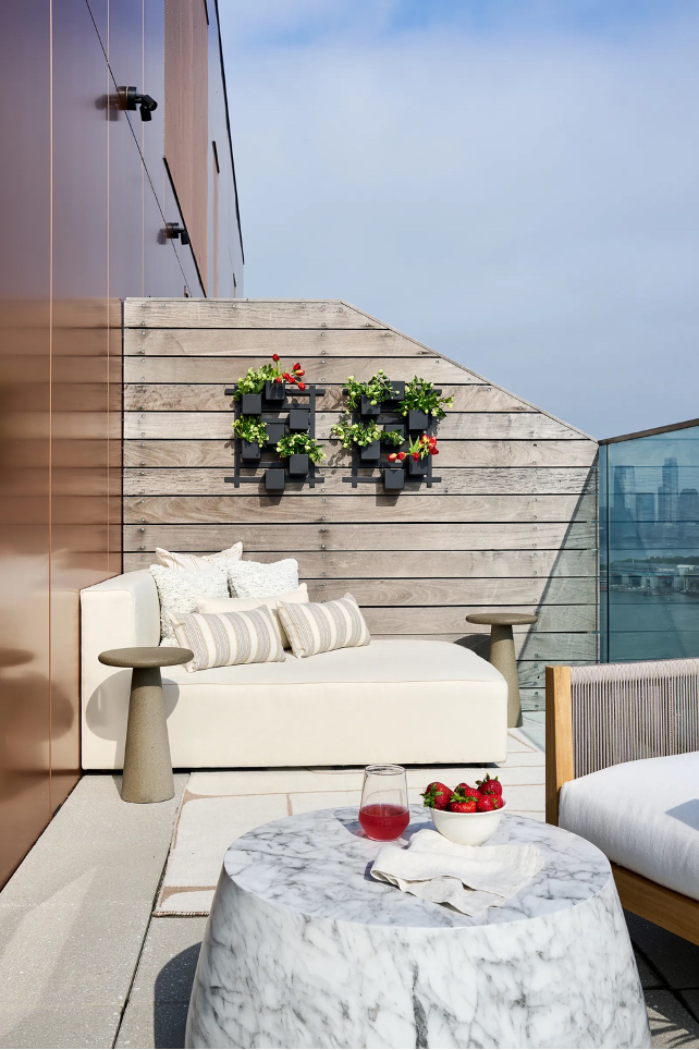 Corner seating on balcony with city and water views. Hanging greenery and flowers on wooden privacy wall, marble side table with bowl of strawberries, linen napkin, and stemless wineglass.