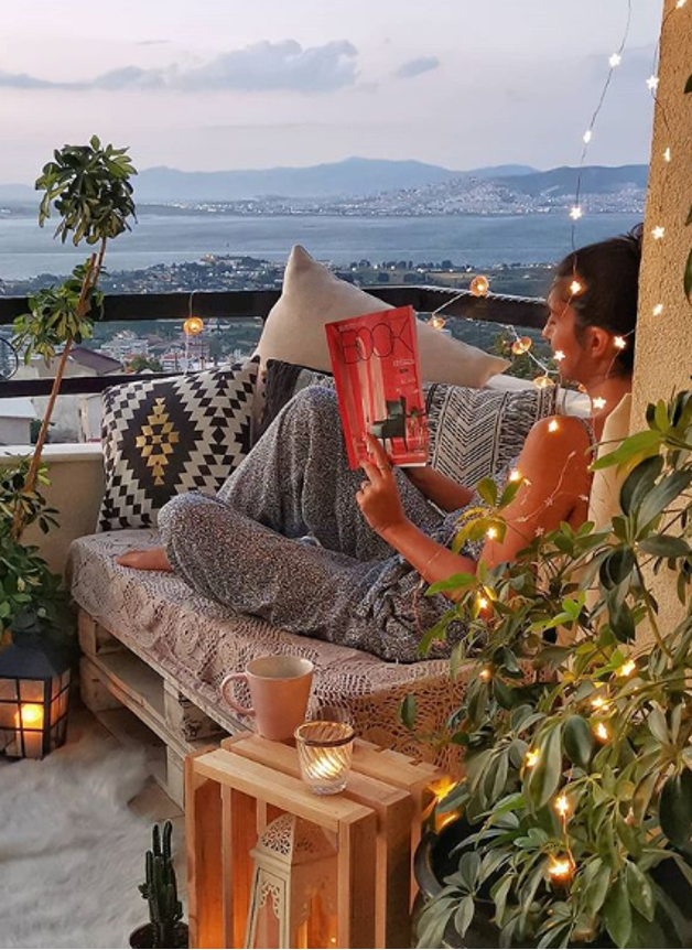 Dreamy oceanfront balcony with greenery, fairy string lights, lanterns, cozy corner seating, and a girl reading a magazine.