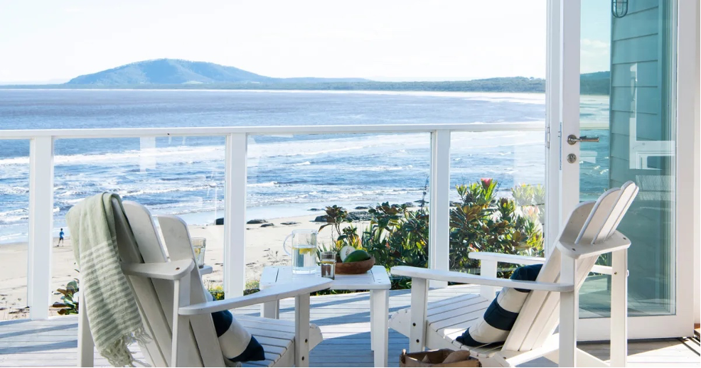 Seaside balcony with Adirondack chairs, striped cushions, and table with water pitcher and wooden bowl with fruit.