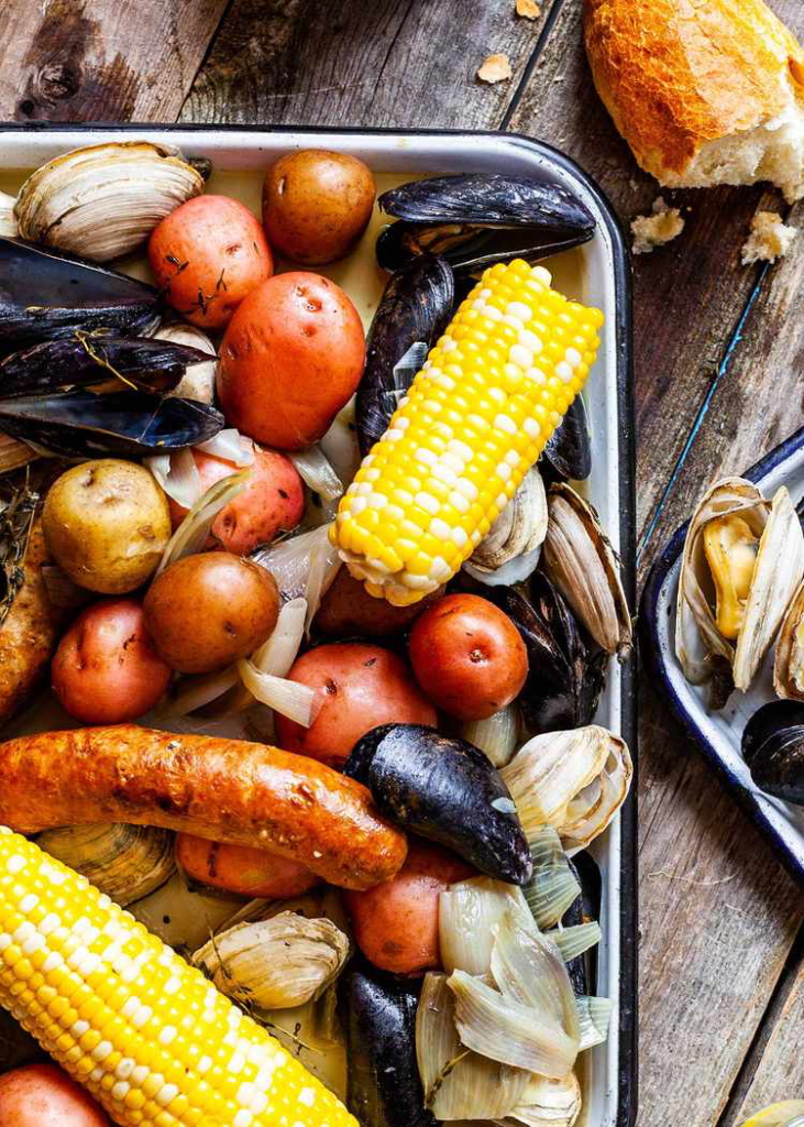 New England Clam Bake served in pan on rustic wooden table with bread and oysters on the side