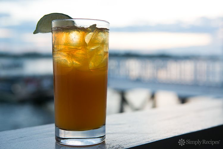 Dark 'n Stormy cocktail set against a sea landscape with a crossing bridge in background. Glass is filled with dark rum, ice cubes, ginger beer, and garnished with a lime wedge.