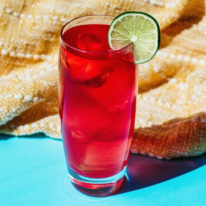 Sea Breeze cocktail in tall glass garnished with a lime wheel and sitting on turquoise blue surface with woven fabric in the background