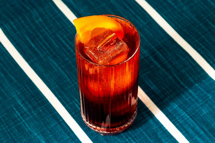 Americano Cocktail in a ridged glass with ice cubes and garnished with an orange slice. Background fabric is deep blue with white stripes.