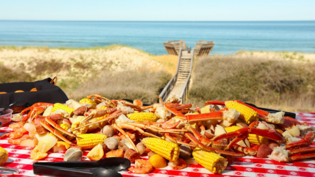 seafood boil on picnic table on the beach featuring crab legs, shrimp, corn, potatoes