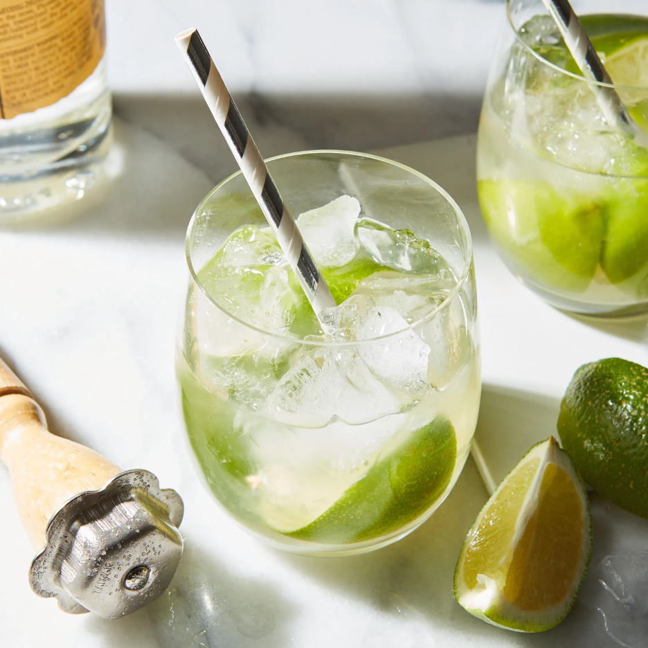 Caipirinha cocktails with limes, ice cubes, a muddler, and striped straws. Cut lime wedges in foreground with a bottle of cachaça in the background.