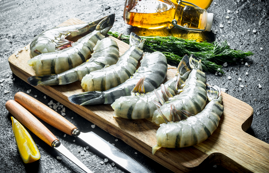 Tiger prawns on wooden cutting board with knives, lemon wedge, fresh herbs, bottled olive oil, and sea salt sprinkles