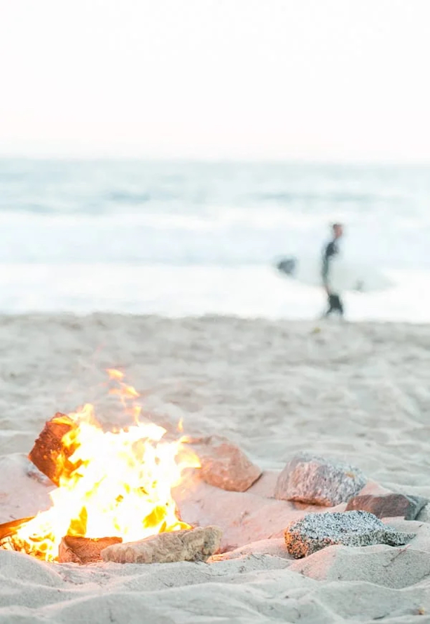 beach bonfire surrounded by stones on white sand with a surfer walking the beach in the background against the ocean waves