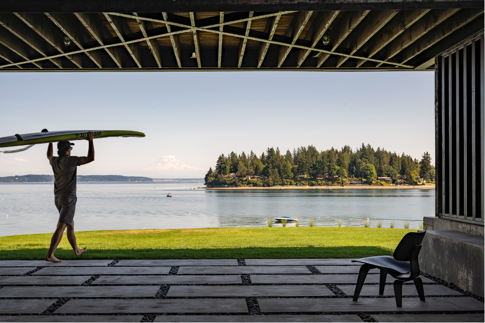 homeowner carrying surfbaord on top of head on the lower level covered beach deck with views of Puget Sound waters and Mount Rainier in backdrop.