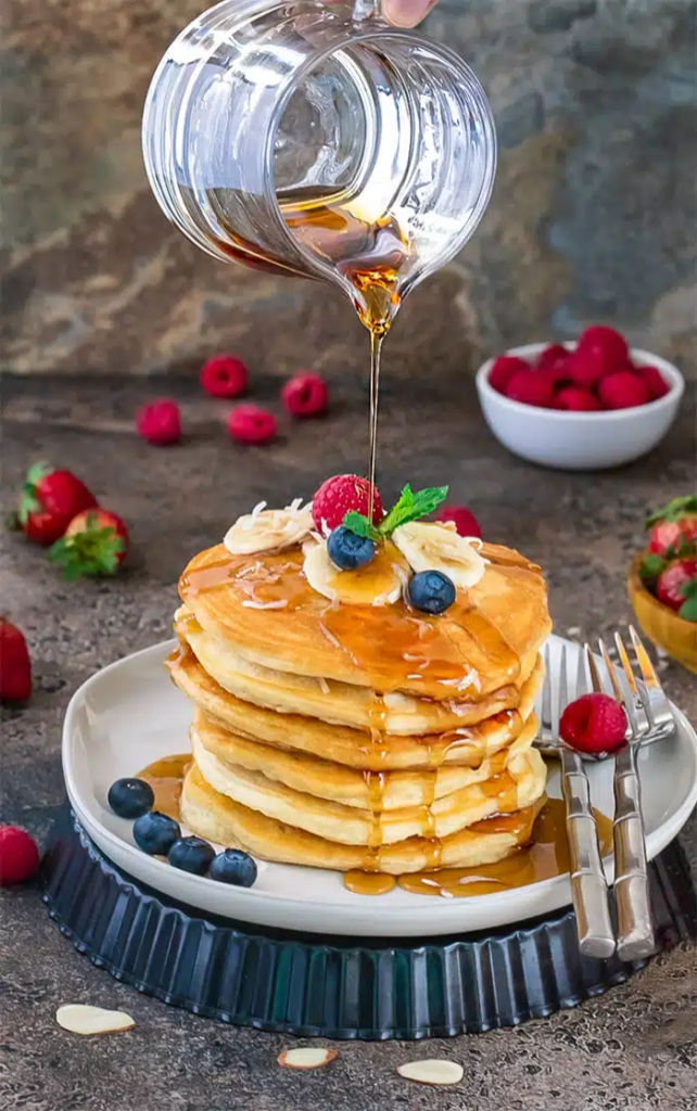 Vegan Coconut Banana Pancakes stacked on a plate and drizzled with syrup, topped with toasted coconut, bananas, blueberries, and raspberries