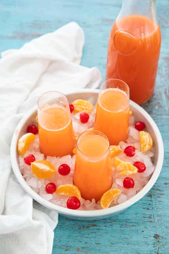 Holiday Mimosa Punch served in stemless glasses in a white tray filled with crushed ice, orange pieces, and marachino cherries.